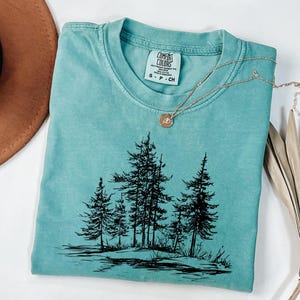 May include: A teal Comfort Colors t-shirt with a black graphic of pine trees. A gold necklace with a small pendant rests on the shirt. The shirt is folded and placed on a white surface. A brown hat and dried leaves are in the background.