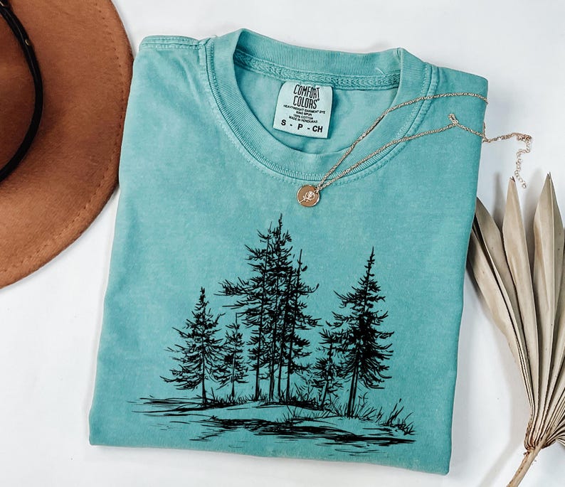 May include: A teal Comfort Colors t-shirt with a black graphic of pine trees. A gold necklace with a small pendant rests on the shirt. The shirt is folded and placed on a white surface. A brown hat and dried leaves are in the background.