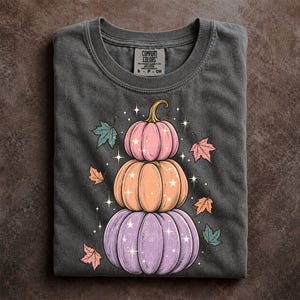 May include: A charcoal gray t-shirt featuring a graphic of three stacked pumpkins in pastel pink, orange, and purple. The design includes scattered autumn leaves and small white starburst accents. The inner neck label reads Comfort Colors.