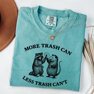 Puede incluir: Camiseta turquesa Comfort Colors con el texto "MORE TRASH CAN LESS TRASH CAN'T" y un gr&aacute;fico de un mapache y una zarig&uuml;eya chocando los cinco. Tambi&eacute;n se ven un collar dorado y un sombrero marr&oacute;n.