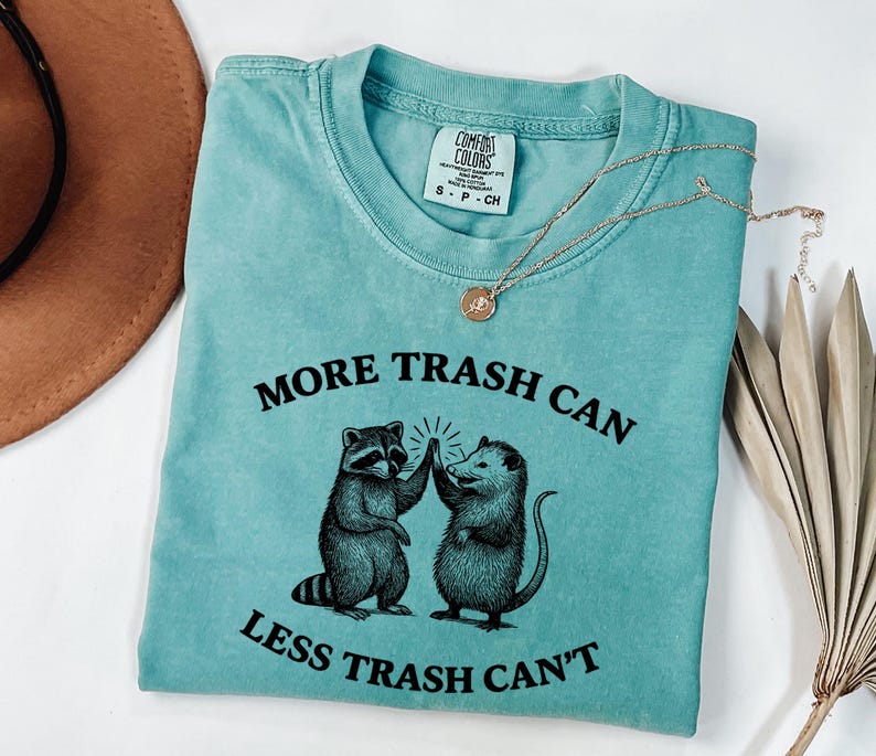 Puede incluir: Camiseta turquesa Comfort Colors con el texto "MORE TRASH CAN LESS TRASH CAN'T" y un gr&aacute;fico de un mapache y una zarig&uuml;eya chocando los cinco. Tambi&eacute;n se ven un collar dorado y un sombrero marr&oacute;n.