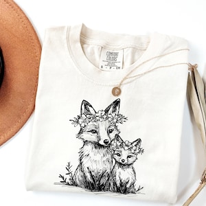 May include: Cream-colored Comfort Colors t-shirt featuring a black and white illustration of two foxes wearing floral crowns. A gold necklace with a pendant rests on the shirt. The shirt has a small tag with the brand name.