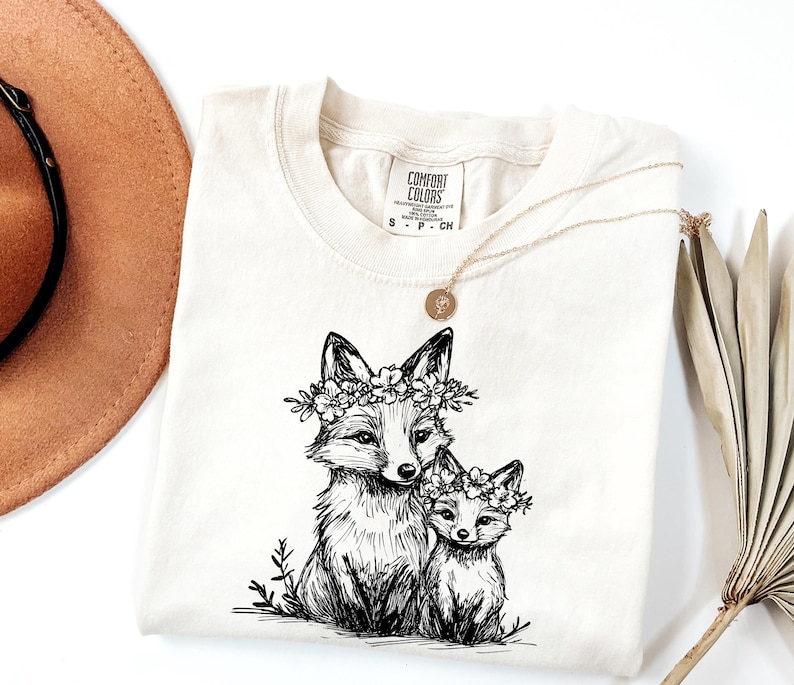 May include: Cream-colored Comfort Colors t-shirt featuring a black and white illustration of two foxes wearing floral crowns. A gold necklace with a pendant rests on the shirt. The shirt has a small tag with the brand name.