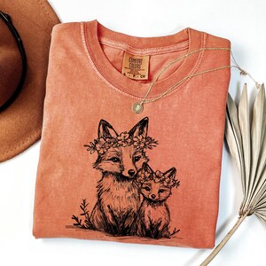 May include: Coral-colored Comfort Colors t-shirt featuring a black illustration of two foxes wearing floral crowns. A gold necklace with a pendant rests on the shirt. A brown hat and dried palm leaf are in the background.