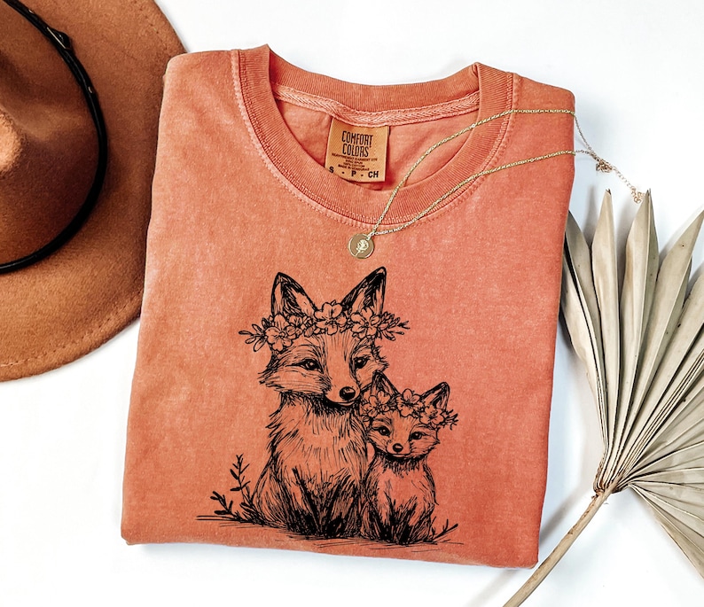 May include: Coral-colored Comfort Colors t-shirt featuring a black illustration of two foxes wearing floral crowns. A gold necklace with a pendant rests on the shirt. A brown hat and dried palm leaf are in the background.