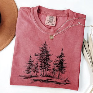 May include: A red-orange Comfort Colors t-shirt with a crew neck and short sleeves. The shirt features a black graphic of several pine trees. A silver necklace with a small pendant is draped across the shirt.