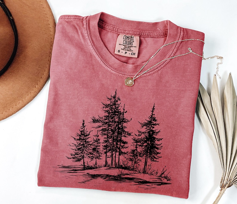 May include: A red-orange Comfort Colors t-shirt with a crew neck and short sleeves. The shirt features a black graphic of several pine trees. A silver necklace with a small pendant is draped across the shirt.