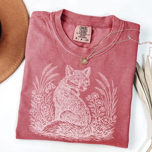 May include: A red Comfort Colors t-shirt with a white fox graphic surrounded by flowers and wheat. The shirt has a crew neck and a small tag at the neckline. A silver necklace with a small pendant is draped across the shirt.