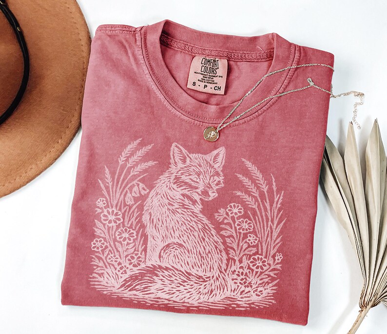 May include: A red Comfort Colors t-shirt with a white fox graphic surrounded by flowers and wheat. The shirt has a crew neck and a small tag at the neckline. A silver necklace with a small pendant is draped across the shirt.
