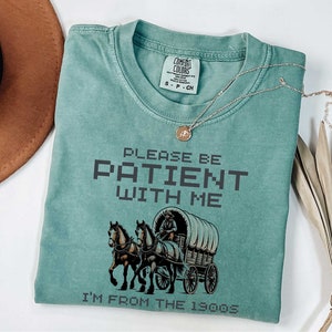 May include: A teal blue t-shirt with a vintage-style illustration of a horse-drawn wagon. The text "PLEASE BE PATIENT WITH ME I'M FROM THE 1900S" is printed in black on the shirt.