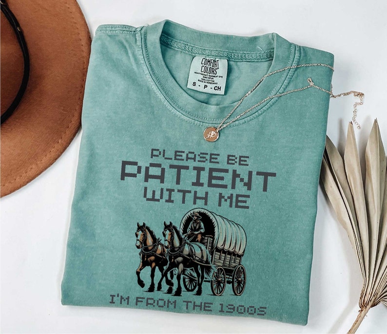 May include: A teal blue t-shirt with a vintage-style illustration of a horse-drawn wagon. The text "PLEASE BE PATIENT WITH ME I'M FROM THE 1900S" is printed in black on the shirt.