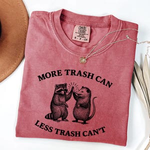 Puede incluir: Una camiseta rosa desgastada con el texto "MORE TRASH CAN LESS TRASH CAN'T" y un gr&aacute;fico de un mapache y una zarig&uuml;eya chocando los cinco. La camiseta tiene cuello redondo y mangas cortas. Una cadena de oro est&aacute; sobre la camiseta.