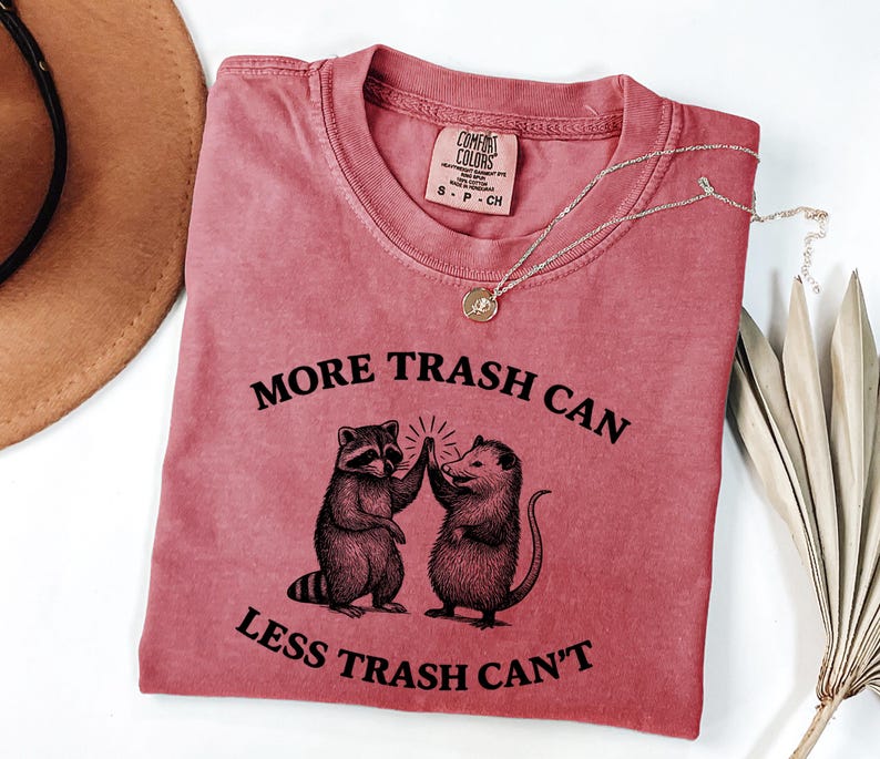 Puede incluir: Una camiseta rosa desgastada con el texto "MORE TRASH CAN LESS TRASH CAN'T" y un gr&aacute;fico de un mapache y una zarig&uuml;eya chocando los cinco. La camiseta tiene cuello redondo y mangas cortas. Una cadena de oro est&aacute; sobre la camiseta.