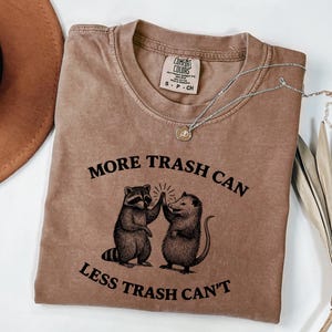 Puede incluir: Camiseta marr&oacute;n con el texto "MORE TRASH CAN LESS TRASH CAN'T" y un gr&aacute;fico de un mapache y una zarig&uuml;eya chocando los cinco. Un collar plateado con un peque&ntilde;o colgante est&aacute; sobre la camiseta.