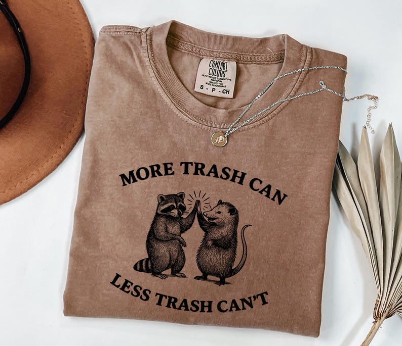 Puede incluir: Camiseta marr&oacute;n con el texto "MORE TRASH CAN LESS TRASH CAN'T" y un gr&aacute;fico de un mapache y una zarig&uuml;eya chocando los cinco. Un collar plateado con un peque&ntilde;o colgante est&aacute; sobre la camiseta.