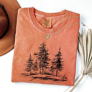 May include: A coral-colored t-shirt featuring a black graphic of pine trees. The shirt has a crew neck and a small tag on the collar. A gold necklace with a pendant is draped across the shirt. A brown hat and a dried palm leaf are also in the image.