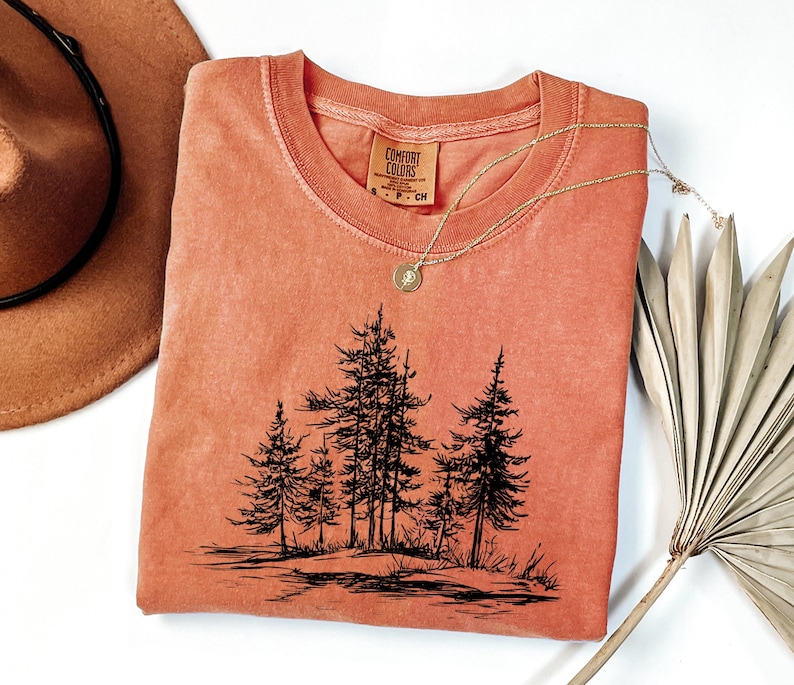 May include: A coral-colored t-shirt featuring a black graphic of pine trees. The shirt has a crew neck and a small tag on the collar. A gold necklace with a pendant is draped across the shirt. A brown hat and a dried palm leaf are also in the image.