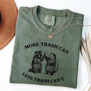 Puede incluir: Camiseta verde oliva con el texto "MORE TRASH CAN LESS TRASH CAN'T" y un gr&aacute;fico de un mapache y una zarig&uuml;eya chocando los cinco. Un collar dorado con un colgante est&aacute; sobre la camiseta.
