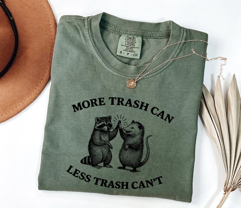 Puede incluir: Camiseta verde oliva con el texto "MORE TRASH CAN LESS TRASH CAN'T" y un gr&aacute;fico de un mapache y una zarig&uuml;eya chocando los cinco. Un collar dorado con un colgante est&aacute; sobre la camiseta.