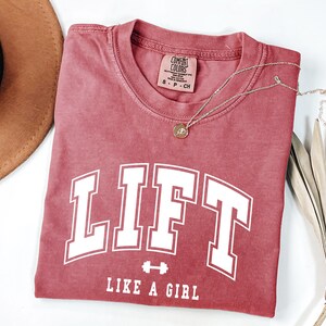 Pode incluir: Uma camiseta vermelha com a palavra "LIFT" em letras brancas grandes, com um gr&aacute;fico de halteres e a frase "LIKE A GIRL" abaixo. Um colar de ouro com um pingente est&aacute; drapeado sobre a camisa.