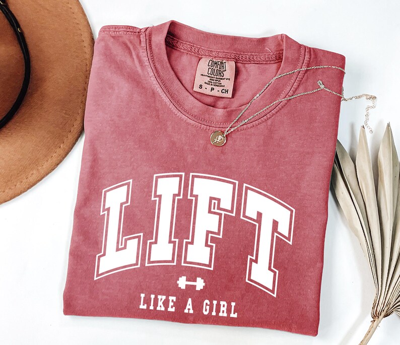 Pode incluir: Uma camiseta vermelha com a palavra "LIFT" em letras brancas grandes, com um gr&aacute;fico de halteres e a frase "LIKE A GIRL" abaixo. Um colar de ouro com um pingente est&aacute; drapeado sobre a camisa.