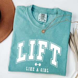 Pode incluir: Camiseta azul-turquesa com a palavra "LIFT" em letras brancas, um gr&aacute;fico de haltere e a frase "LIKE A GIRL". Um colar de ouro com um pingente repousa sobre a camisa. A camisa est&aacute; dobrada, com um chap&eacute;u marrom e folhas de palmeira secas no fundo.