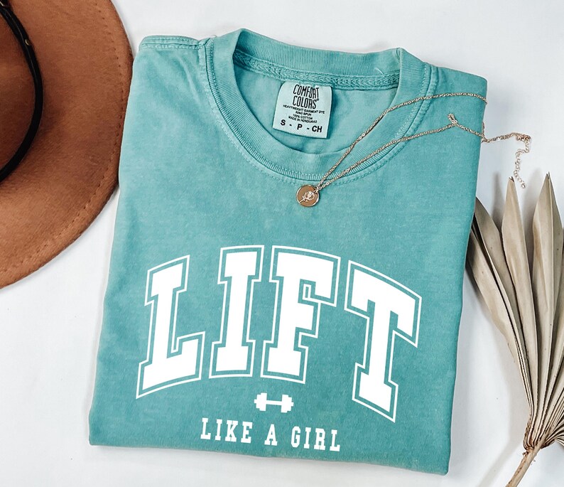 Pode incluir: Camiseta azul-turquesa com a palavra "LIFT" em letras brancas, um gr&aacute;fico de haltere e a frase "LIKE A GIRL". Um colar de ouro com um pingente repousa sobre a camisa. A camisa est&aacute; dobrada, com um chap&eacute;u marrom e folhas de palmeira secas no fundo.