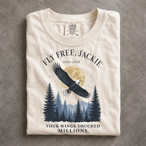 May include: A cream-colored crew neck t-shirt featuring a graphic of a bald eagle flying over a forest of pine trees and a full moon. The text reads FLY FREE, JACKIE 2012-2026 and YOUR WINGS TOUCHED MILLIONS. The shirt has a Comfort Colors brand label.