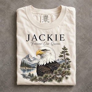 May include: A cream-colored t-shirt featuring a graphic of a bald eagle, snow-capped mountains, a lake, and pine branches. The text reads JACKIE Forever Our Queen in black lettering. The shirt has a Comfort Colors brand label at the inner neckline.