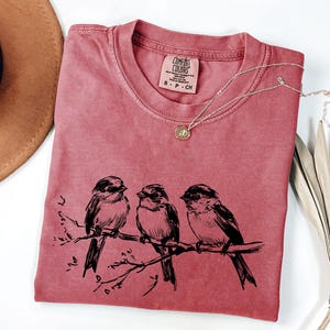 May include: A dusty rose Comfort Colors t-shirt featuring a black illustration of three birds perched on a branch. A delicate silver necklace with a small pendant rests on the shirt. The shirt has a small Comfort Colors tag.