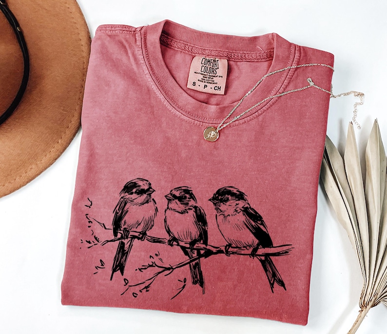 May include: A dusty rose Comfort Colors t-shirt featuring a black illustration of three birds perched on a branch. A delicate silver necklace with a small pendant rests on the shirt. The shirt has a small Comfort Colors tag.
