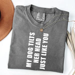 May include: A gray Comfort Colors t-shirt with the text "MY DOG TILTS HER HEAD JUST LIKE YOU" in white. A gold necklace with a pendant is on the shirt. The shirt is folded on a white surface.
