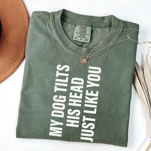 以下が含まれることがあります： 「MY DOG TILTS HIS HEAD JUST LIKE YOU」の文字が白で縦にプリントされたオリーブグリーンのTシャツ。ペンダント付きのゴールドネックレスがシャツの上にあります。シャツは白い面に折りたたまれています。