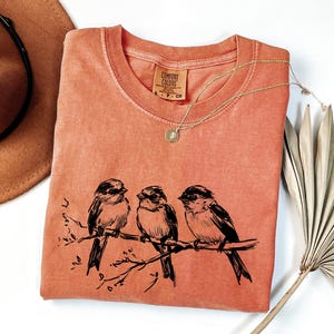 May include: Coral-colored t-shirt with a crew neck and a black illustration of three birds perched on a branch. A gold necklace with a pendant is draped across the shirt. The shirt has a Comfort Colors tag.