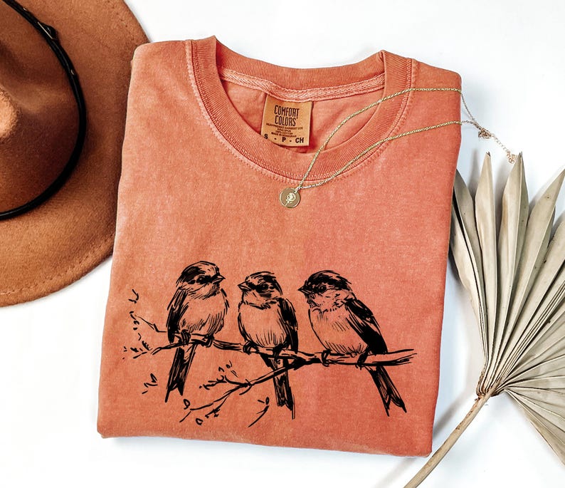 May include: Coral-colored t-shirt with a crew neck and a black illustration of three birds perched on a branch. A gold necklace with a pendant is draped across the shirt. The shirt has a Comfort Colors tag.