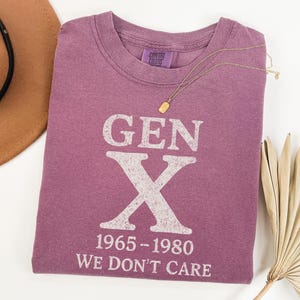 May include: A purple t-shirt with the text "GEN X 1965-1980 WE DON'T CARE" in white. A gold necklace and a brown hat are also in the image. The shirt is made of a soft material.