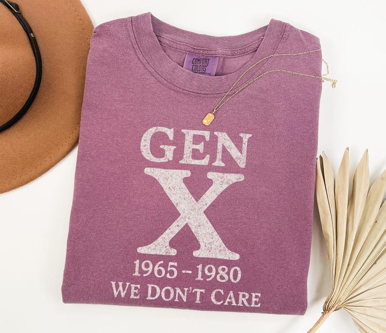 May include: A purple t-shirt with the text "GEN X 1965-1980 WE DON'T CARE" in white. A gold necklace and a brown hat are also in the image. The shirt is made of a soft material.