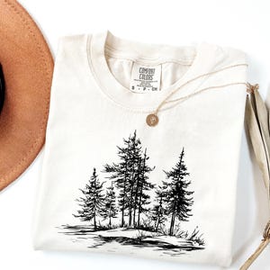 May include: Cream-colored Comfort Colors t-shirt featuring a black and white illustration of pine trees. A gold necklace with a pendant rests on the shirt. A brown hat and dried plant are in the background.