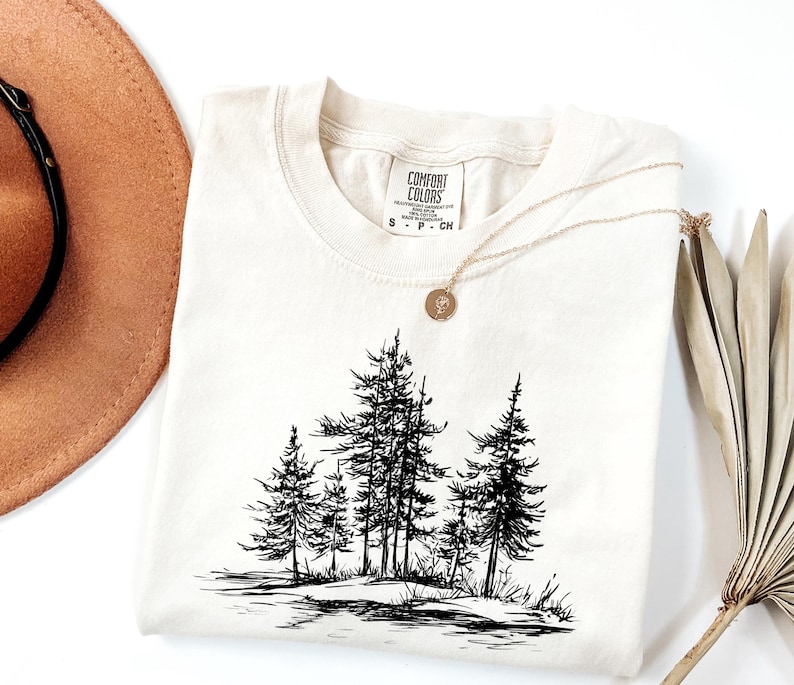 May include: Cream-colored Comfort Colors t-shirt featuring a black and white illustration of pine trees. A gold necklace with a pendant rests on the shirt. A brown hat and dried plant are in the background.