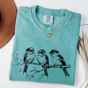 May include: A turquoise Comfort Colors t-shirt with a black illustration of three birds perched on a branch. A gold necklace with a small pendant rests on the shirt. The shirt is folded, with the tag visible.