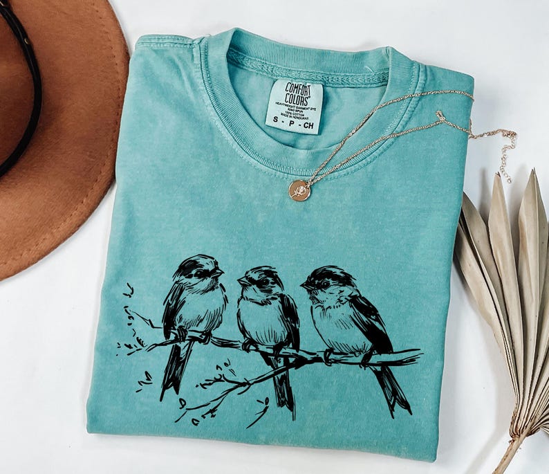 May include: A turquoise Comfort Colors t-shirt with a black illustration of three birds perched on a branch. A gold necklace with a small pendant rests on the shirt. The shirt is folded, with the tag visible.