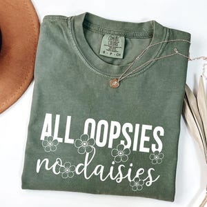 May include: An olive green t-shirt with the phrase "ALL OOPSIES no daisies" in white script and floral accents. A gold necklace with a pendant rests on the shirt. The shirt is folded and displayed with a brown hat and dried plant.