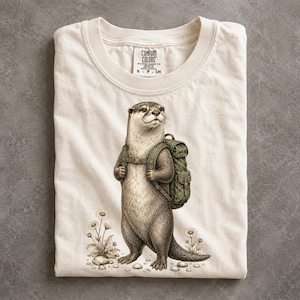 May include: A cream-colored t-shirt featuring a graphic illustration of an otter wearing a green backpack. The otter is standing upright, surrounded by small white flowers. The inner neck label displays the text Comfort Colors and size S CH.