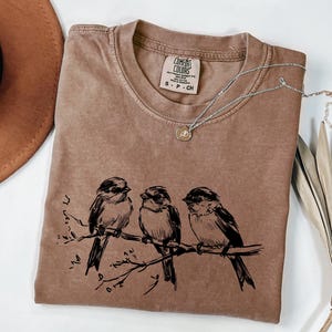 May include: A brown Comfort Colors t-shirt with a crew neck, featuring a black illustration of three birds perched on a branch. A silver necklace with a small pendant rests on the shirt. The shirt is size S-P-CH.