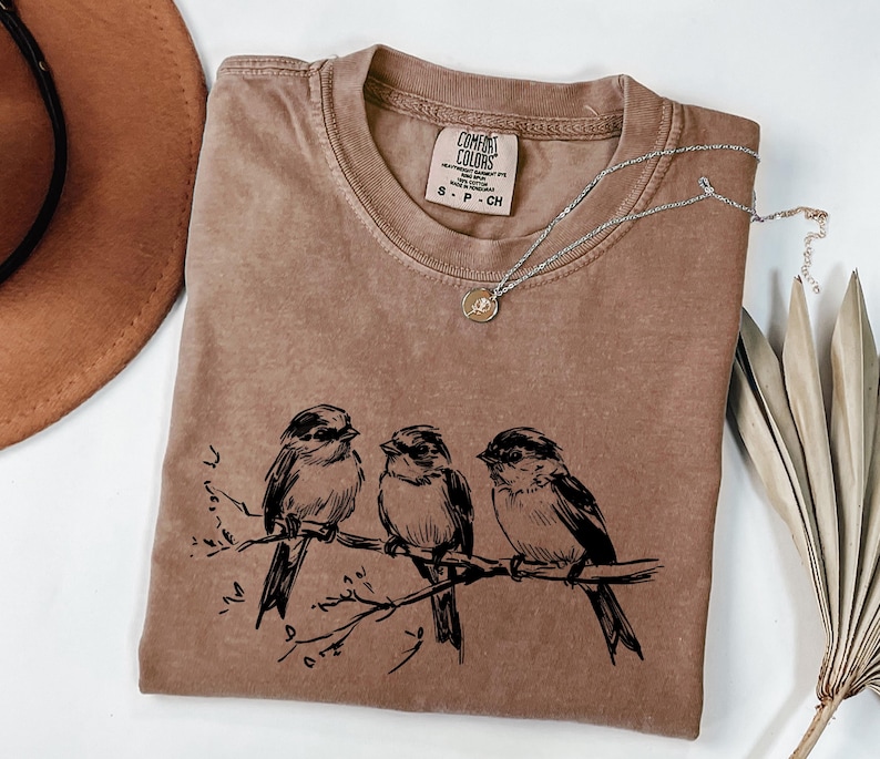 May include: A brown Comfort Colors t-shirt with a crew neck, featuring a black illustration of three birds perched on a branch. A silver necklace with a small pendant rests on the shirt. The shirt is size S-P-CH.