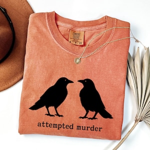 May include: Coral-colored t-shirt featuring two black crow silhouettes facing each other. The phrase "attempted murder" is printed below the birds. A gold necklace with a pendant is draped across the shirt.