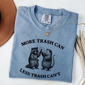Puede incluir: Camiseta azul claro con el texto "MORE TRASH CAN LESS TRASH CAN'T" y un gr&aacute;fico de un mapache y una zarig&uuml;eya chocando los cinco. Un collar dorado con un colgante est&aacute; sobre la camiseta.