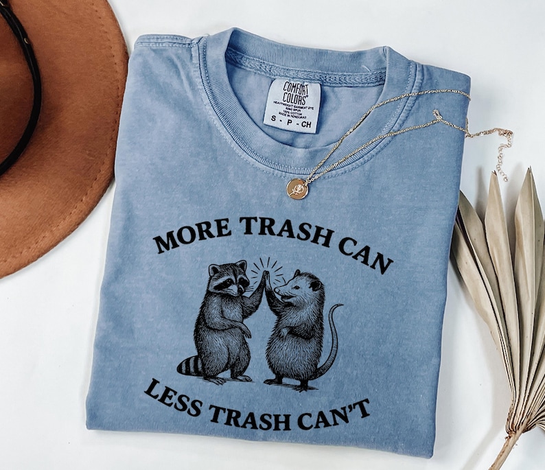 Puede incluir: Camiseta azul claro con el texto "MORE TRASH CAN LESS TRASH CAN'T" y un gr&aacute;fico de un mapache y una zarig&uuml;eya chocando los cinco. Un collar dorado con un colgante est&aacute; sobre la camiseta.