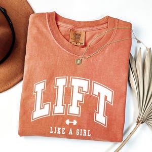 Pode incluir: Uma camiseta cor de coral com a palavra "LIFT" em letras brancas grandes e a frase "LIKE A GIRL" abaixo. Um colar de ouro com um pingente e um chap&eacute;u marrom tamb&eacute;m s&atilde;o vis&iacute;veis.