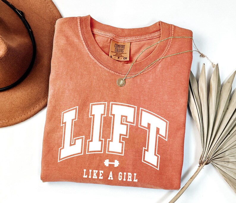 Pode incluir: Uma camiseta cor de coral com a palavra "LIFT" em letras brancas grandes e a frase "LIKE A GIRL" abaixo. Um colar de ouro com um pingente e um chap&eacute;u marrom tamb&eacute;m s&atilde;o vis&iacute;veis.